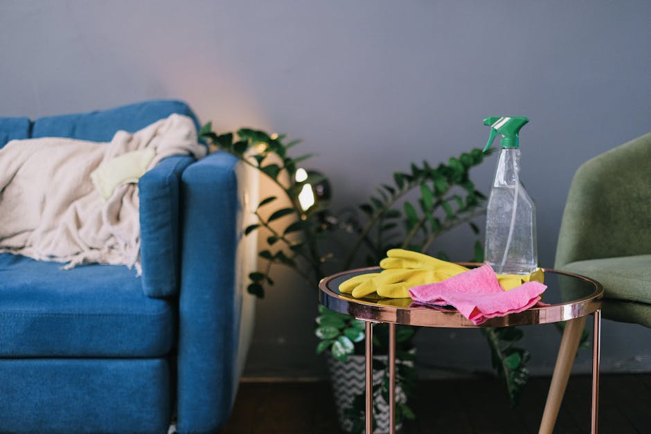 A living room scene featuring a blue fabric sofa with a beige throw blanket draped over one armrest, situated next to a side table. The side table holds cleaning supplies, including yellow rubber glov