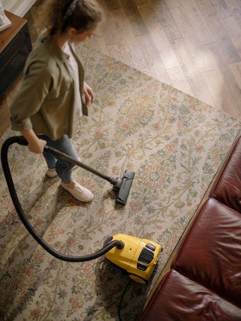 A woman kneeling on a patterned carpet in a domestic setting, operating a yellow and black portable vacuum cleaner with a flexible hose. She is wearing a beige shirt and blue jeans, and is focused on 