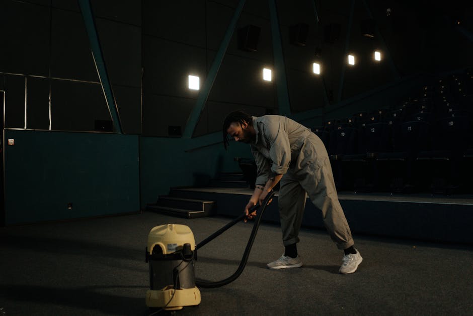 A person dressed in full protective cleaning gear, including a white coverall, mask, gloves, and blue shoe covers, is using a vacuum or extraction machine to deep clean a beige carpet in a bright, mod